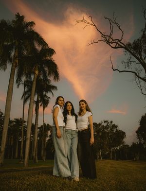 Three women in white tops and jeans pose outdoors among palm trees at sunset with dramatic pink clouds.
