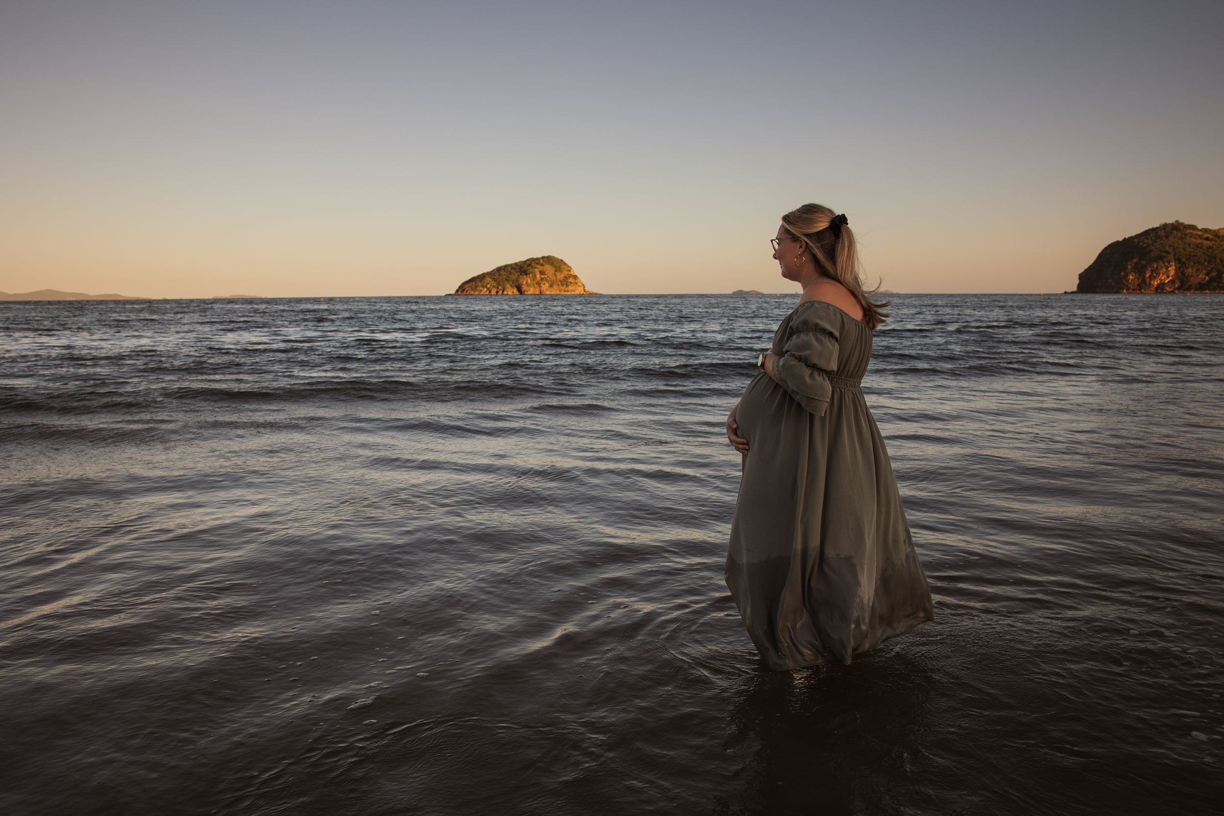 A pregnant woman in a flowing grey dress stands in shallow ocean water at sunset with islands in the background.