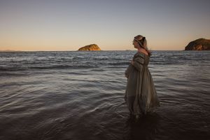 A pregnant woman in a flowing grey dress stands in shallow ocean water at sunset with islands in the background.