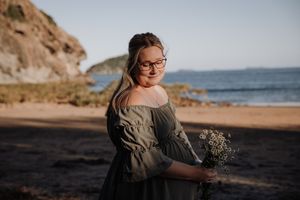 A smiling young woman wearing glasses and a grey off-shoulder dress holds a small bouquet of wildflowers on a beach.