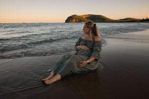 A pregnant woman in a sage green dress sits on a sandy beach at sunset with ocean waves and hills behind her.