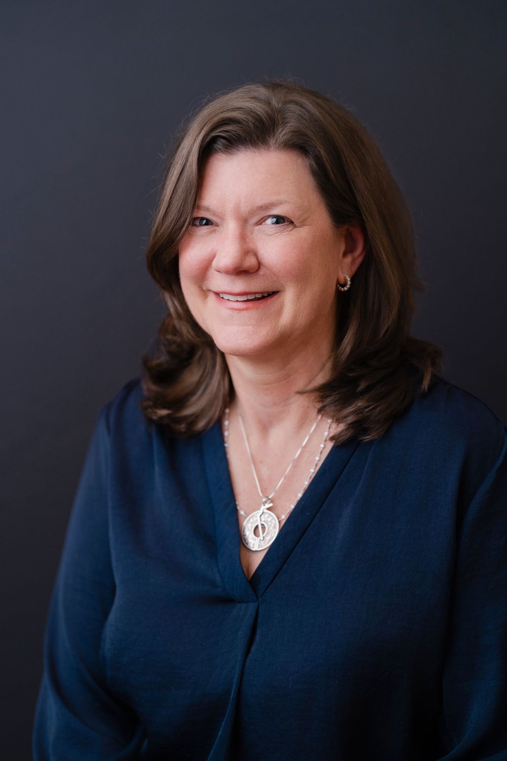 Professional portrait of person wearing navy v-neck sweater with pendant necklace against dark backdrop.