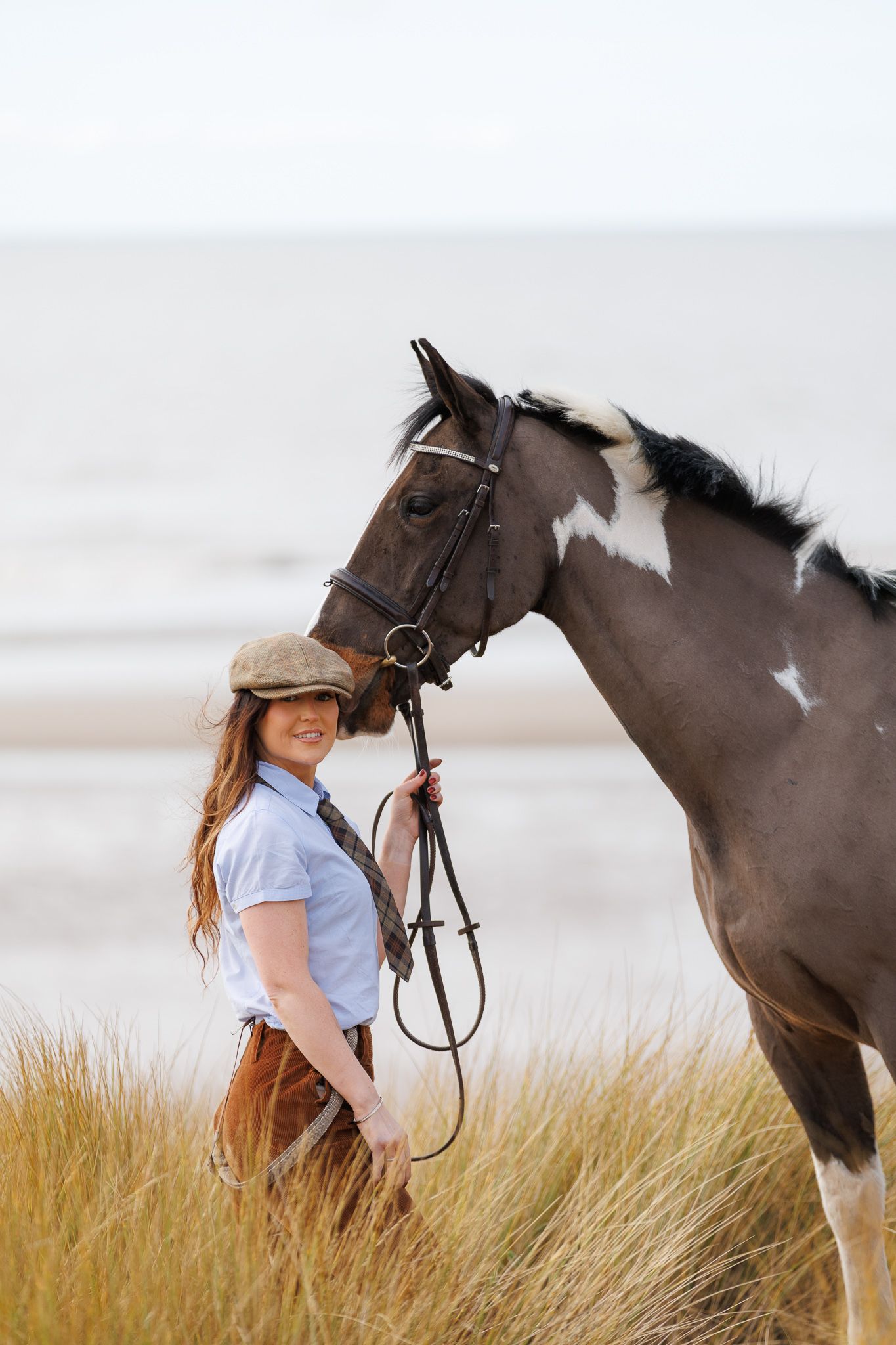 A person in a pink sweater and jeans stands with a black horse against a backdrop of golden autumn foliage.