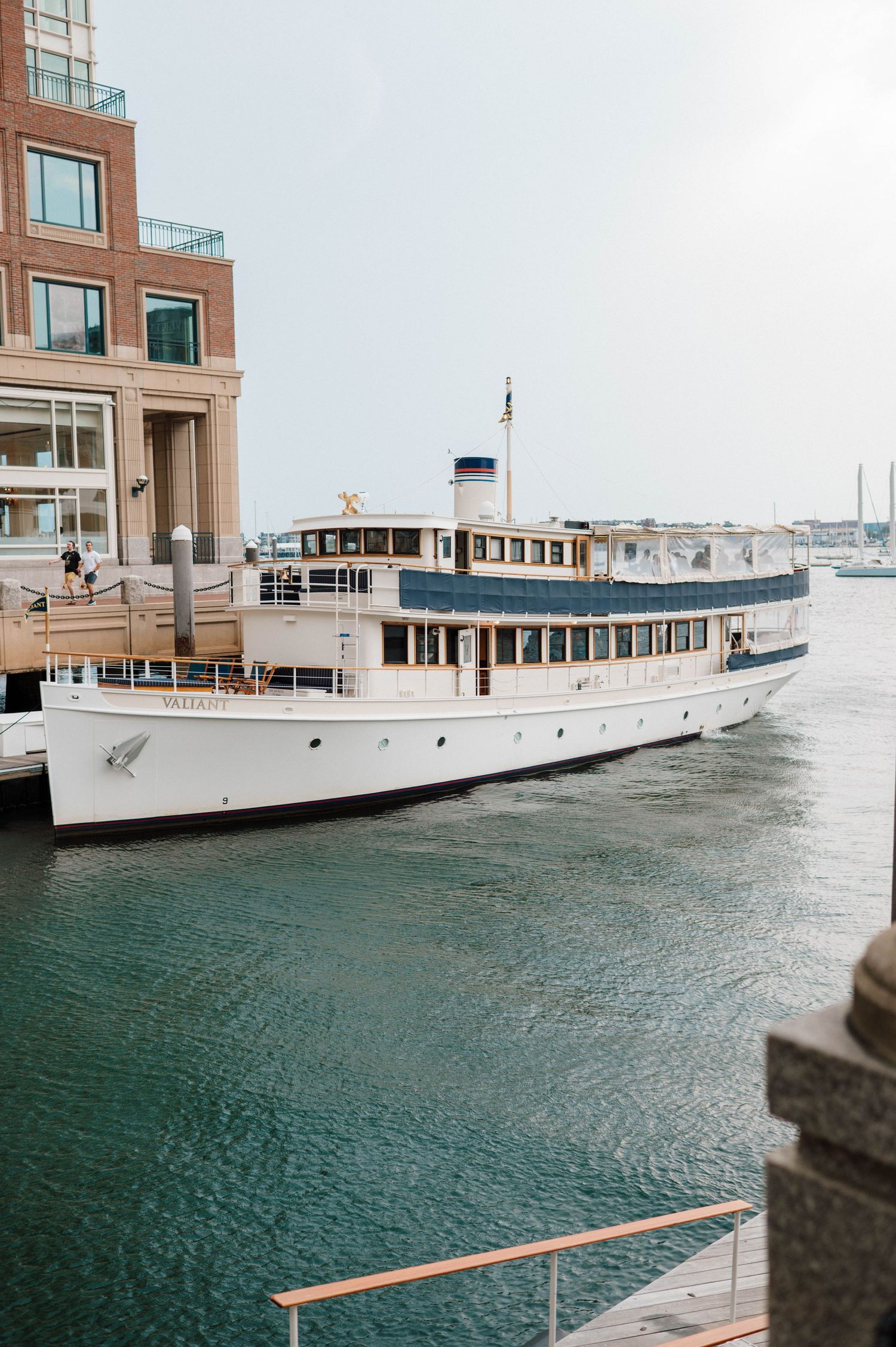 A white vintage boat docked at a modern waterfront building with glass windows and brick facade.