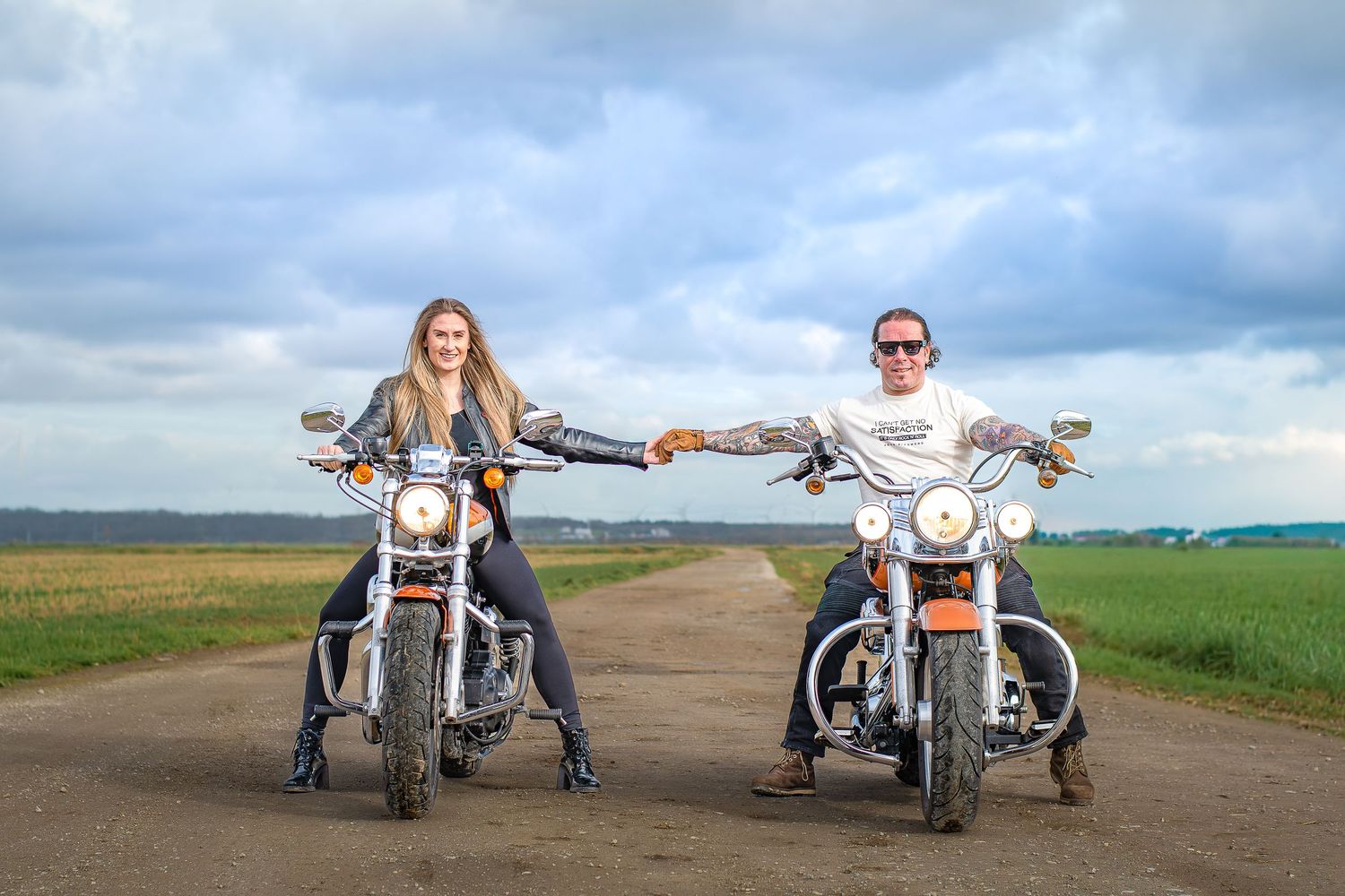 Two people sit on motorcycles while holding hands on a dirt road with dramatic cloudy skies in the background.