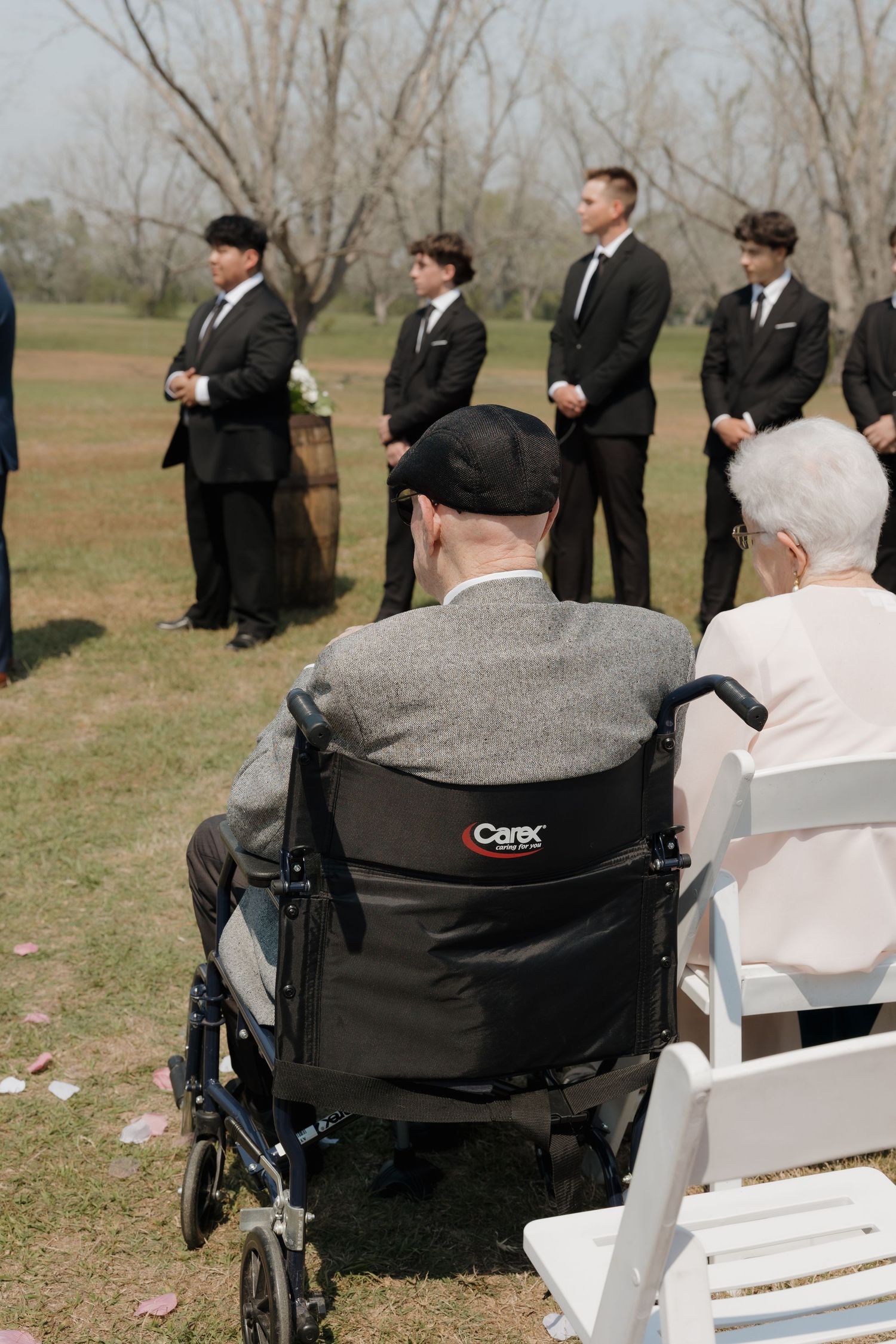 A row of wedding guests in formal attire seated in white chairs outdoors with some using wheelchairs.
