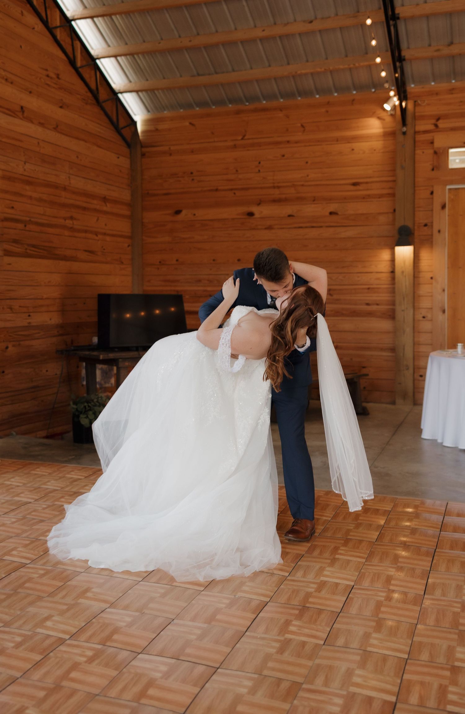 Wedding couple performing dramatic dip dance move on wooden dance floor in rustic venue.