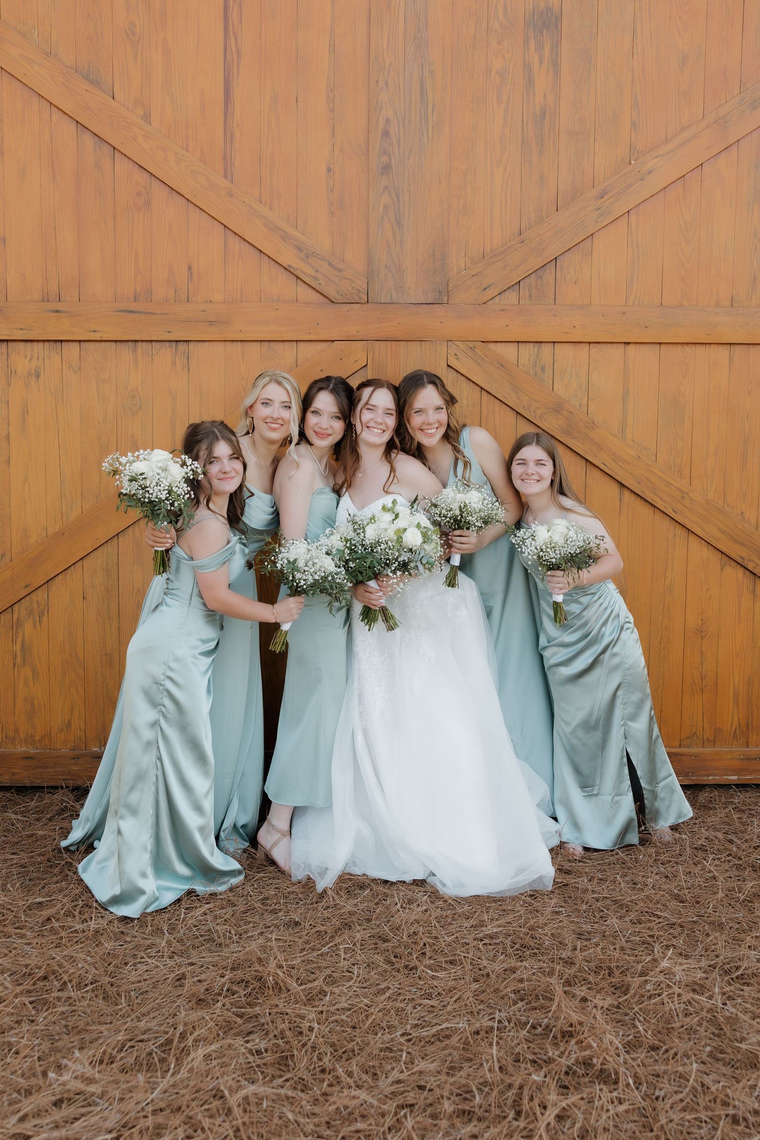 Bridal party in sage green dresses posing together against wooden barn door.