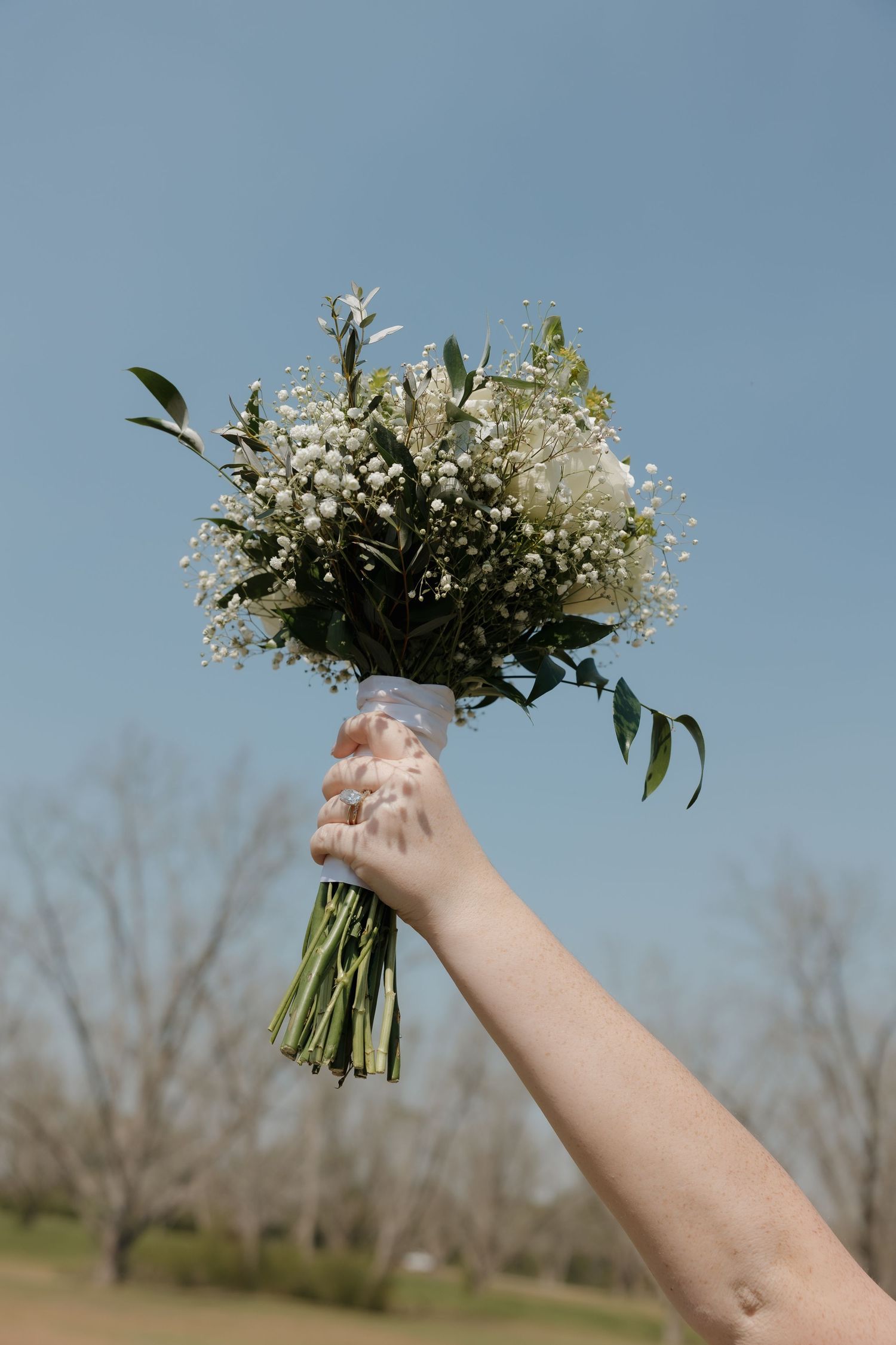 A hand holds up a delicate white floral bouquet with greenery against a blue sky background.