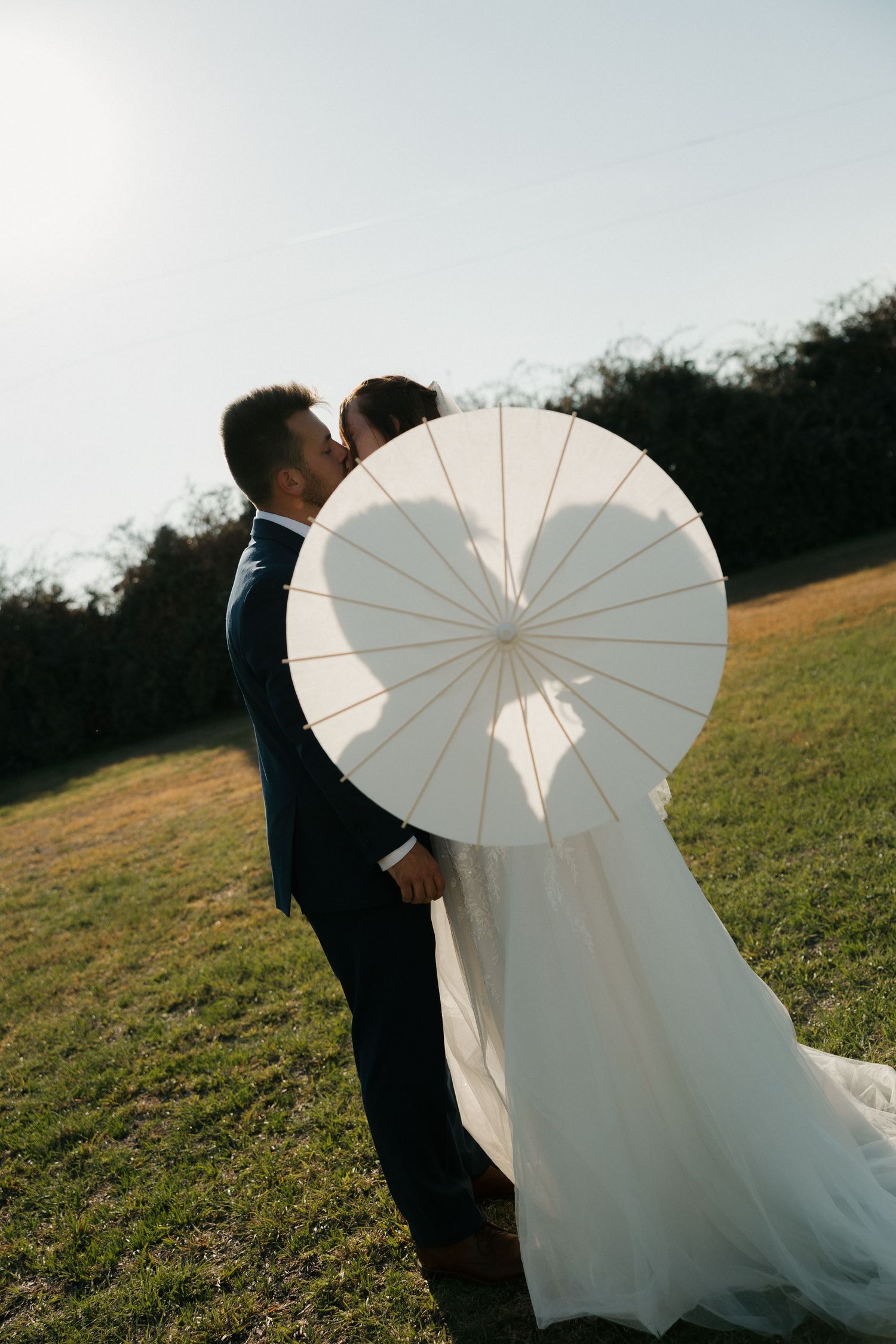Couple with white parasol in outdoor wedding photoshoot on grassy hillside.