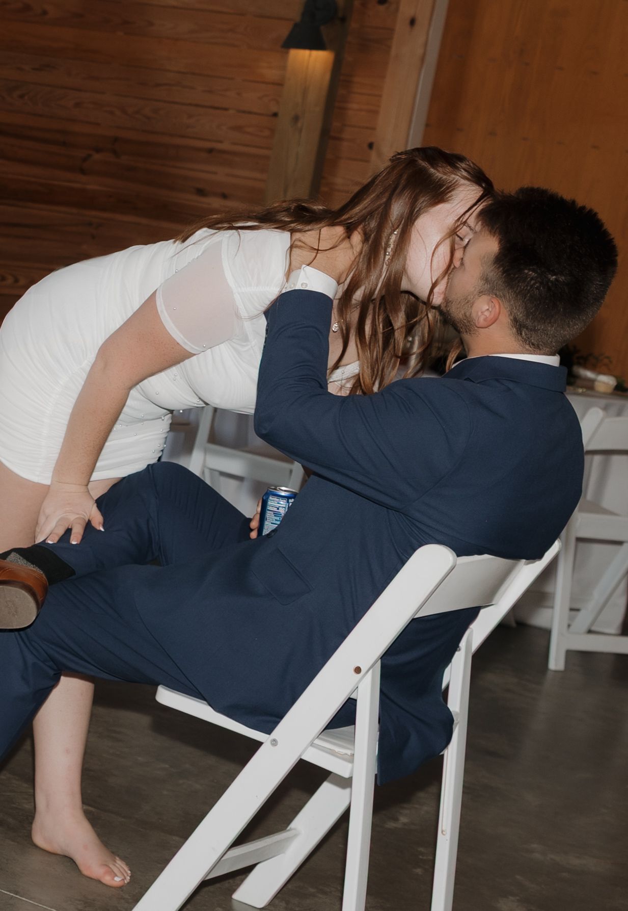 A couple in wedding attire playfully dips backwards in white chairs during a reception celebration.
