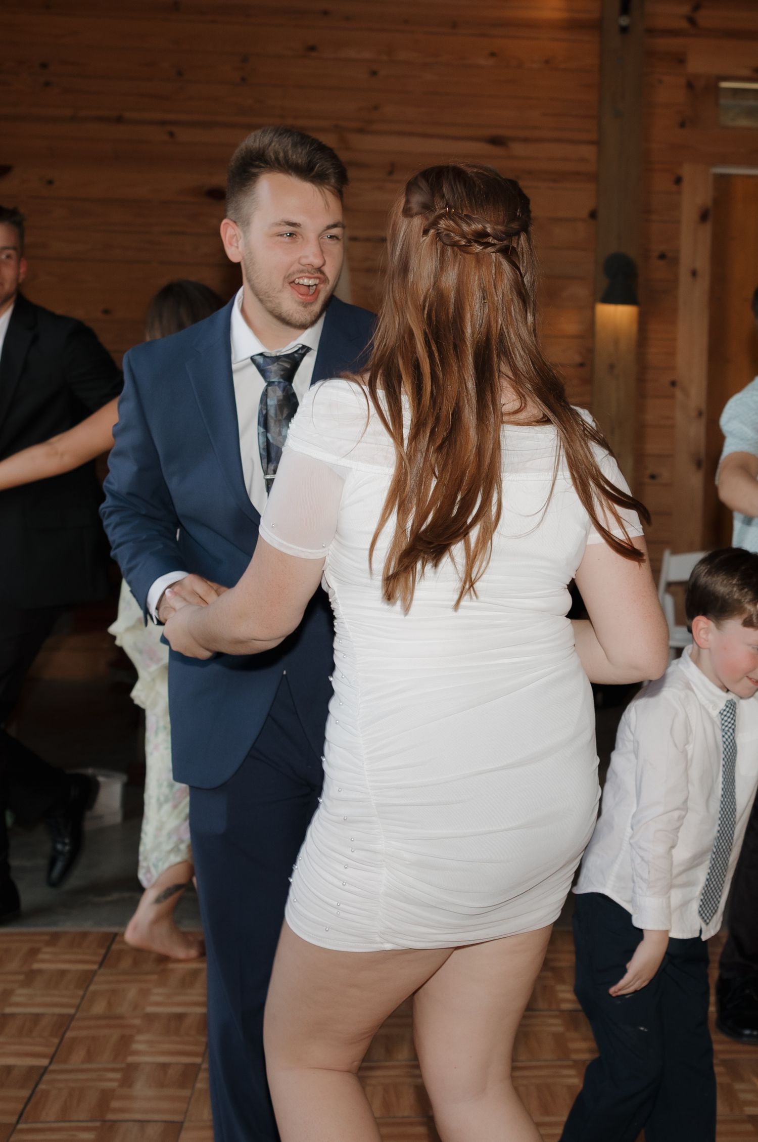 A couple dances together at a wedding reception in a rustic wooden venue, with the bride wearing a short white dress.