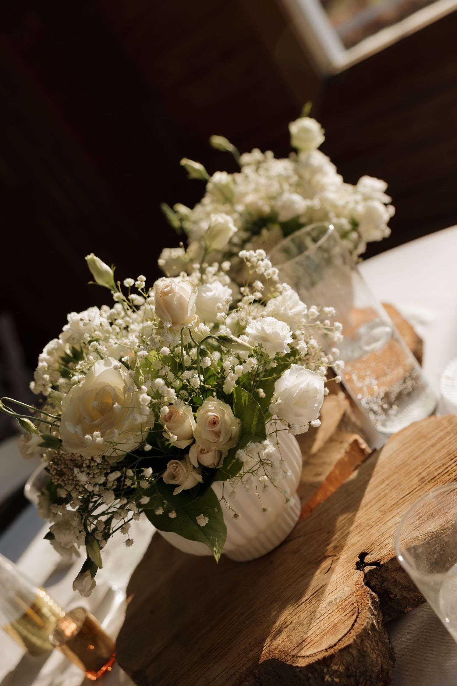 White floral wedding centerpieces with baby's breath and roses arranged on wooden surfaces in a rustic setting.