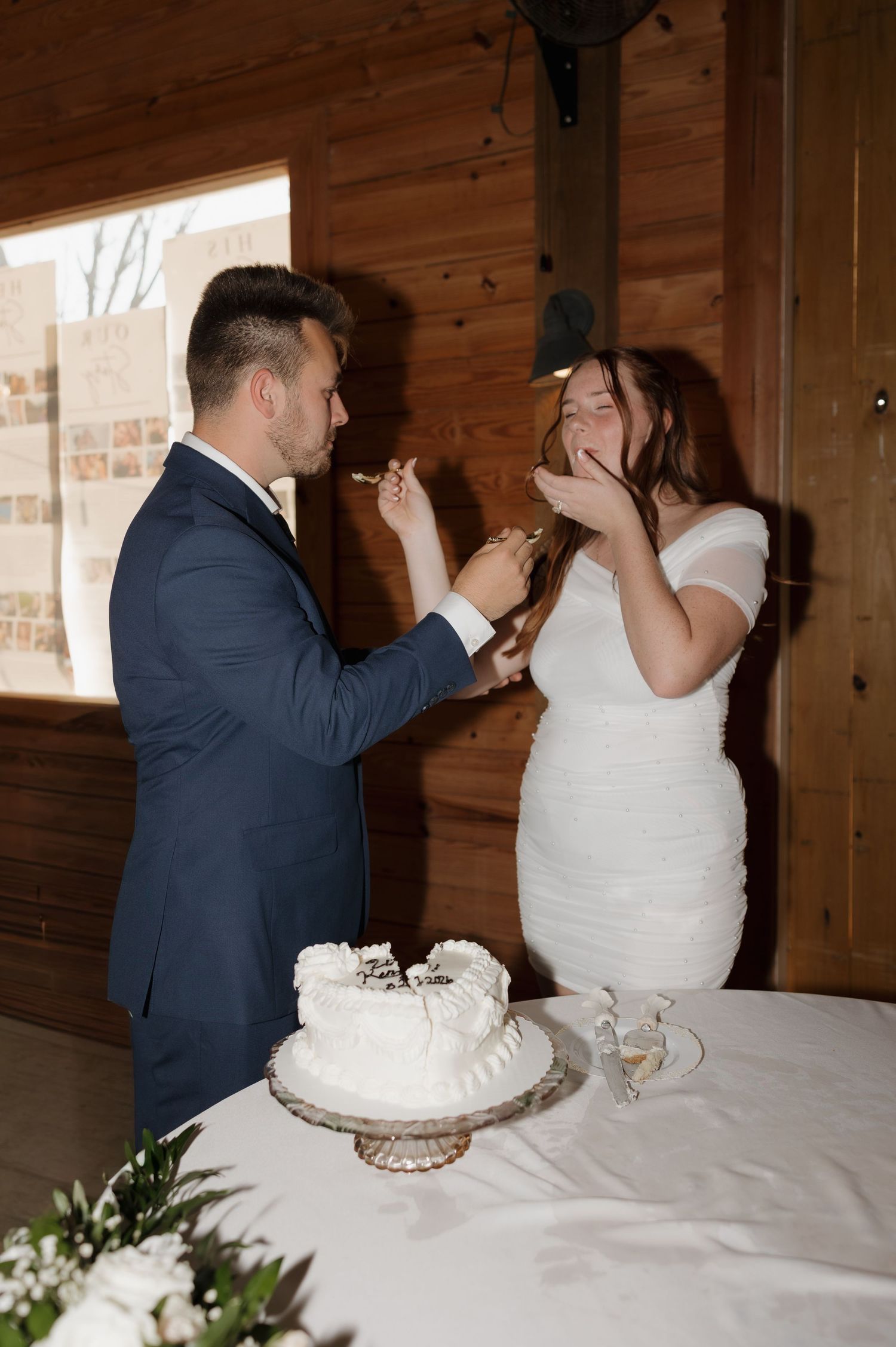 Newlyweds share a playful moment while feeding each other wedding cake at a rustic wooden venue.