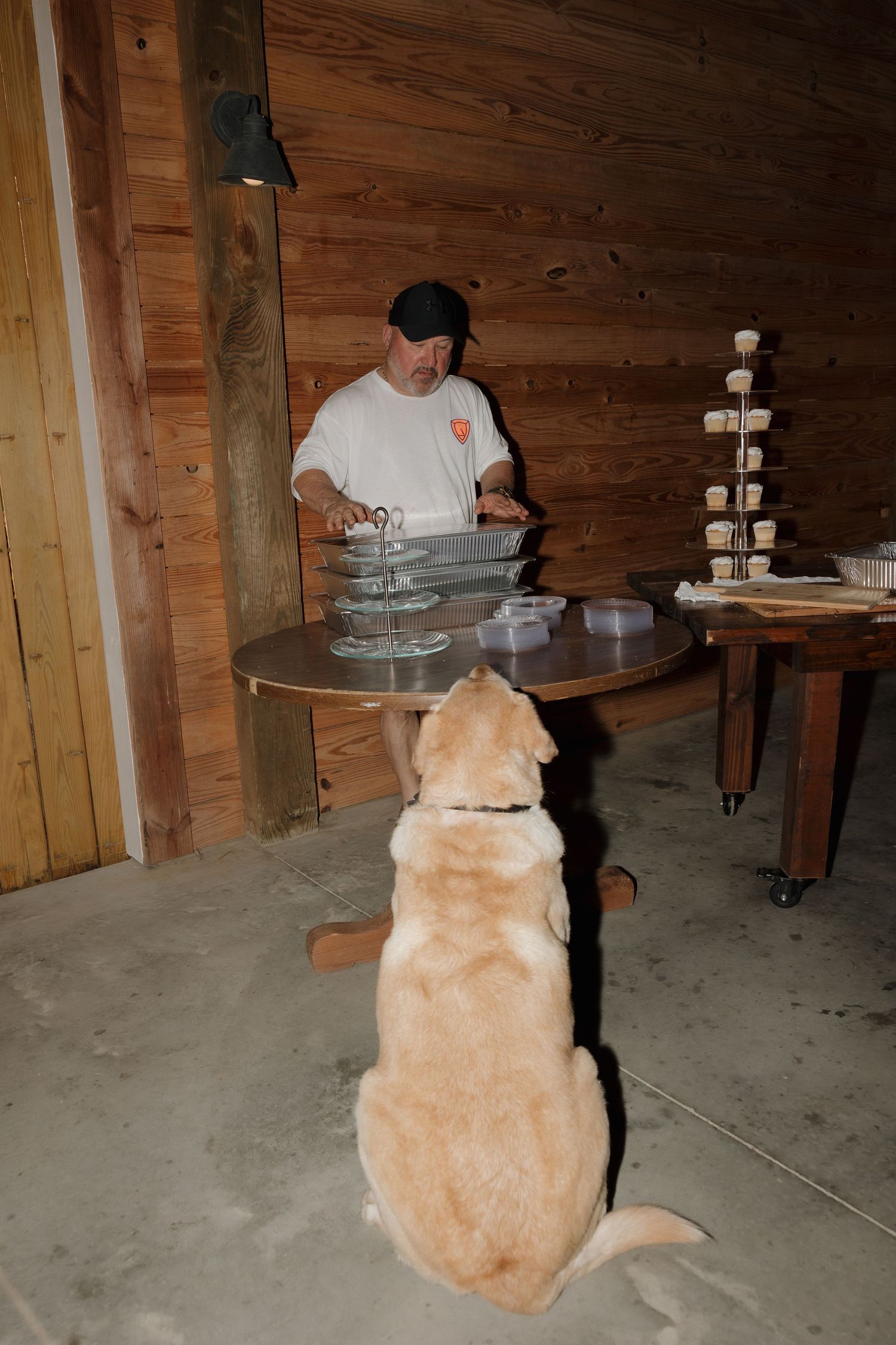 A golden retriever sits patiently watching as someone prepares food at tables in a wooden barn setting.