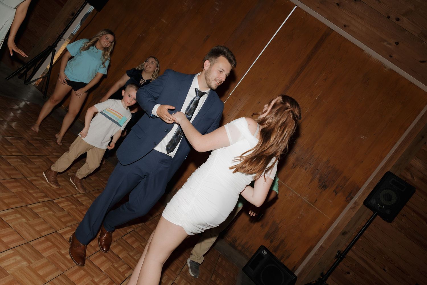 A group of people dancing and celebrating at a rustic indoor wedding reception venue with wooden walls and floors.