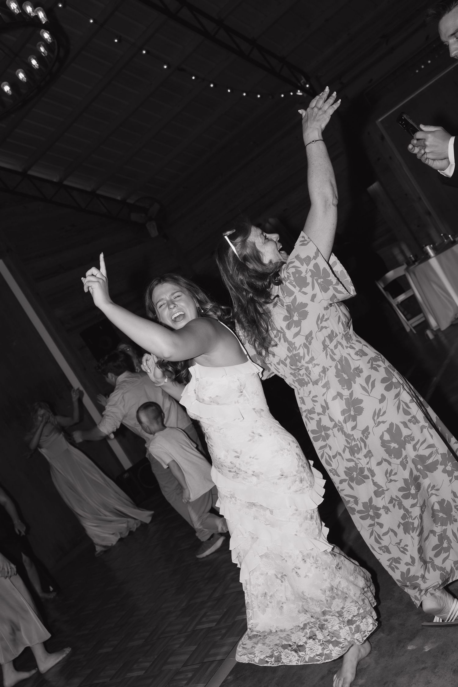 Black and white photo of people dancing energetically at a party in floral dresses with arms raised.