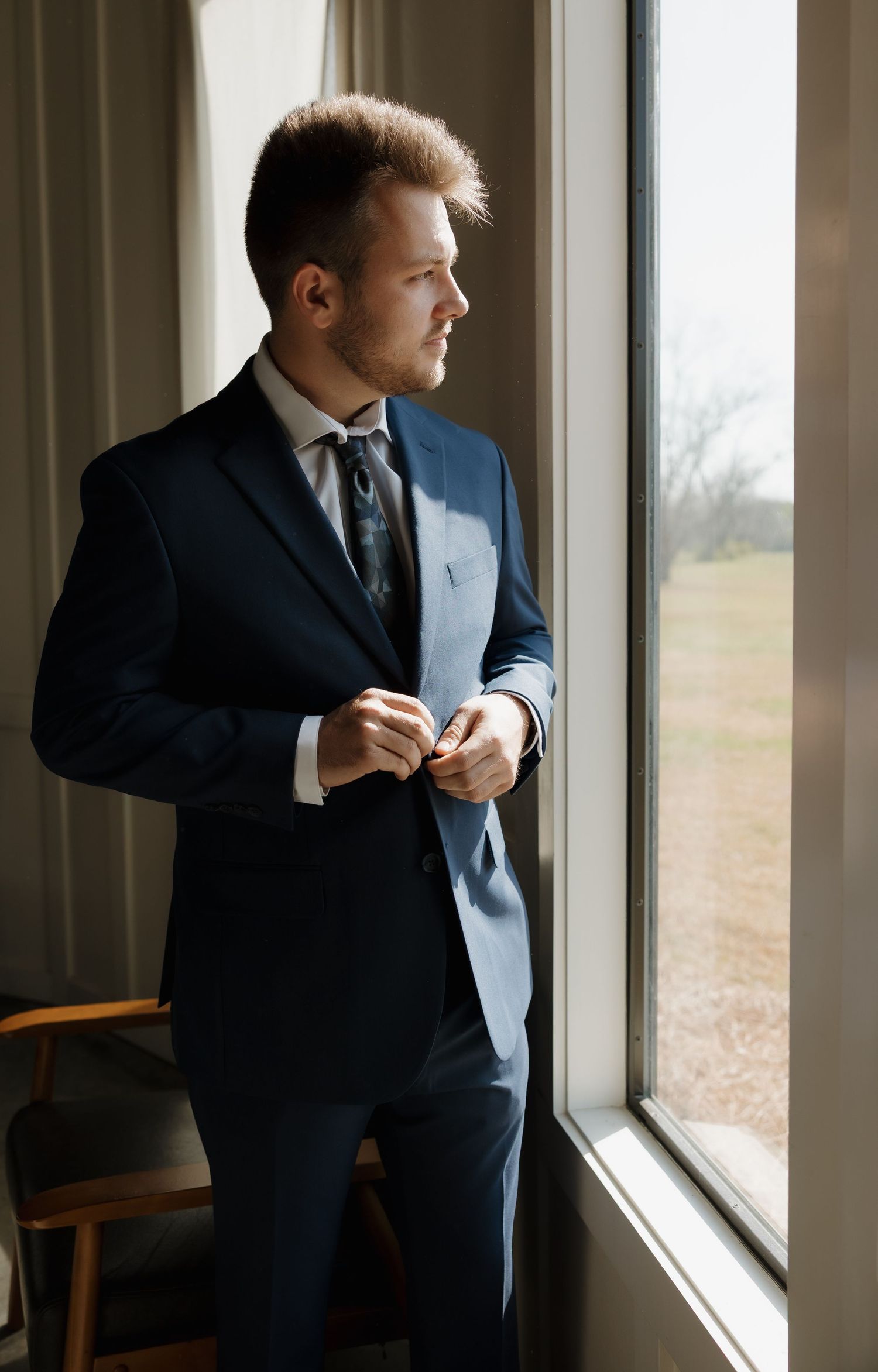 Groom adjusting tie while standing by window in contemplative pre-wedding moment.