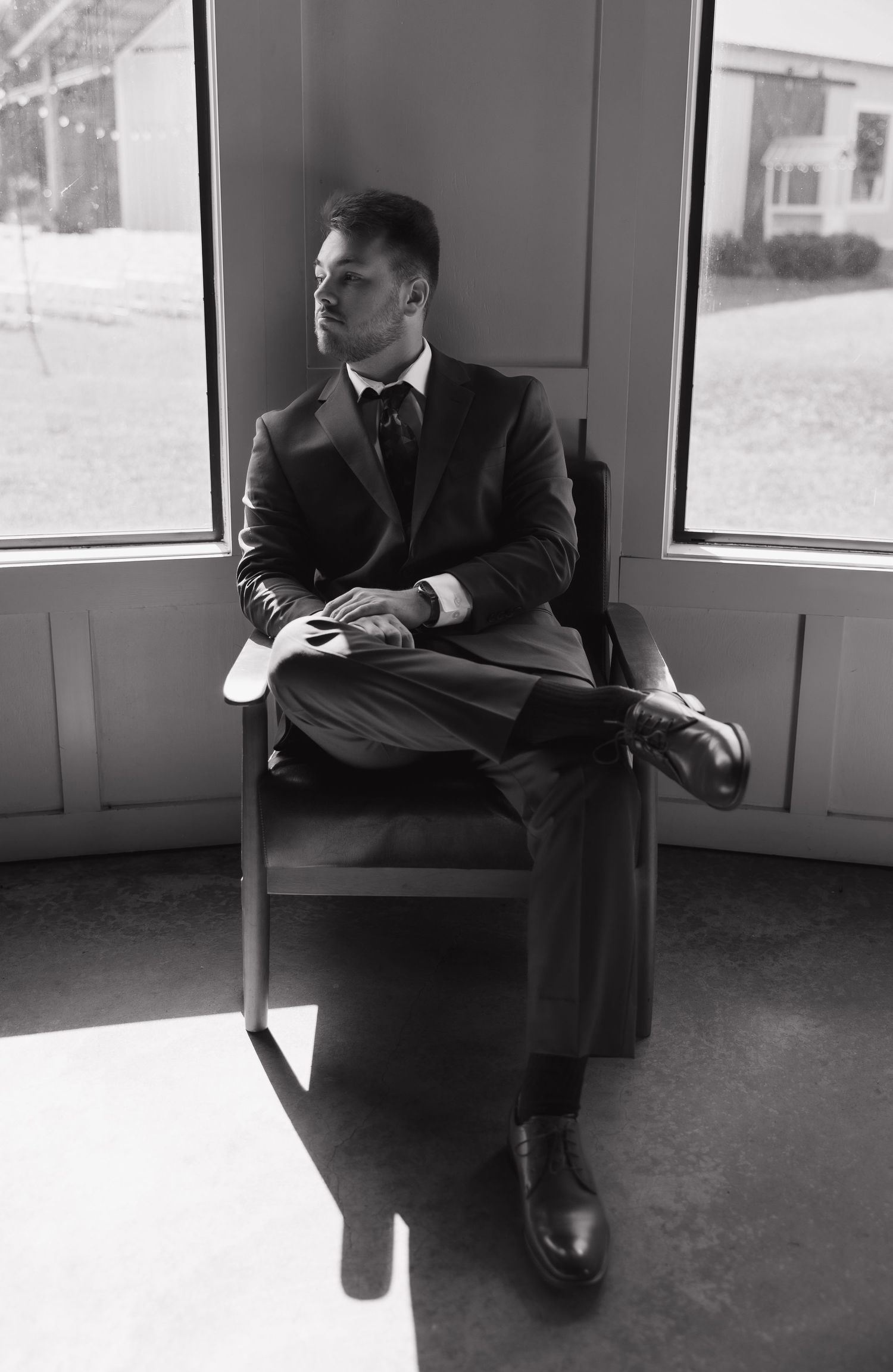 Man in dark suit sitting thoughtfully in empty room with dramatic lighting.