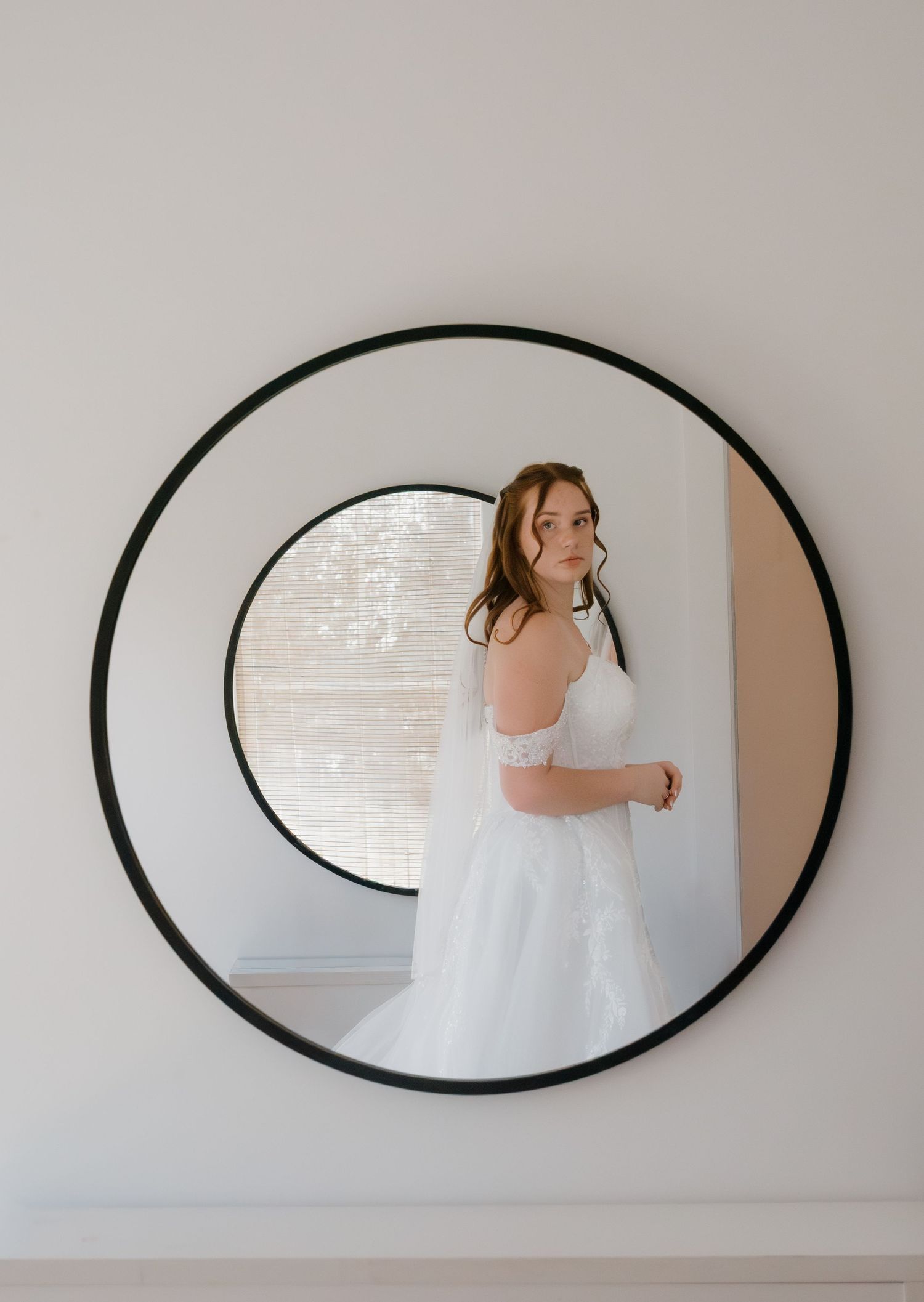 Wedding dress reflection captured in oval mirror mounted on light gray wall.