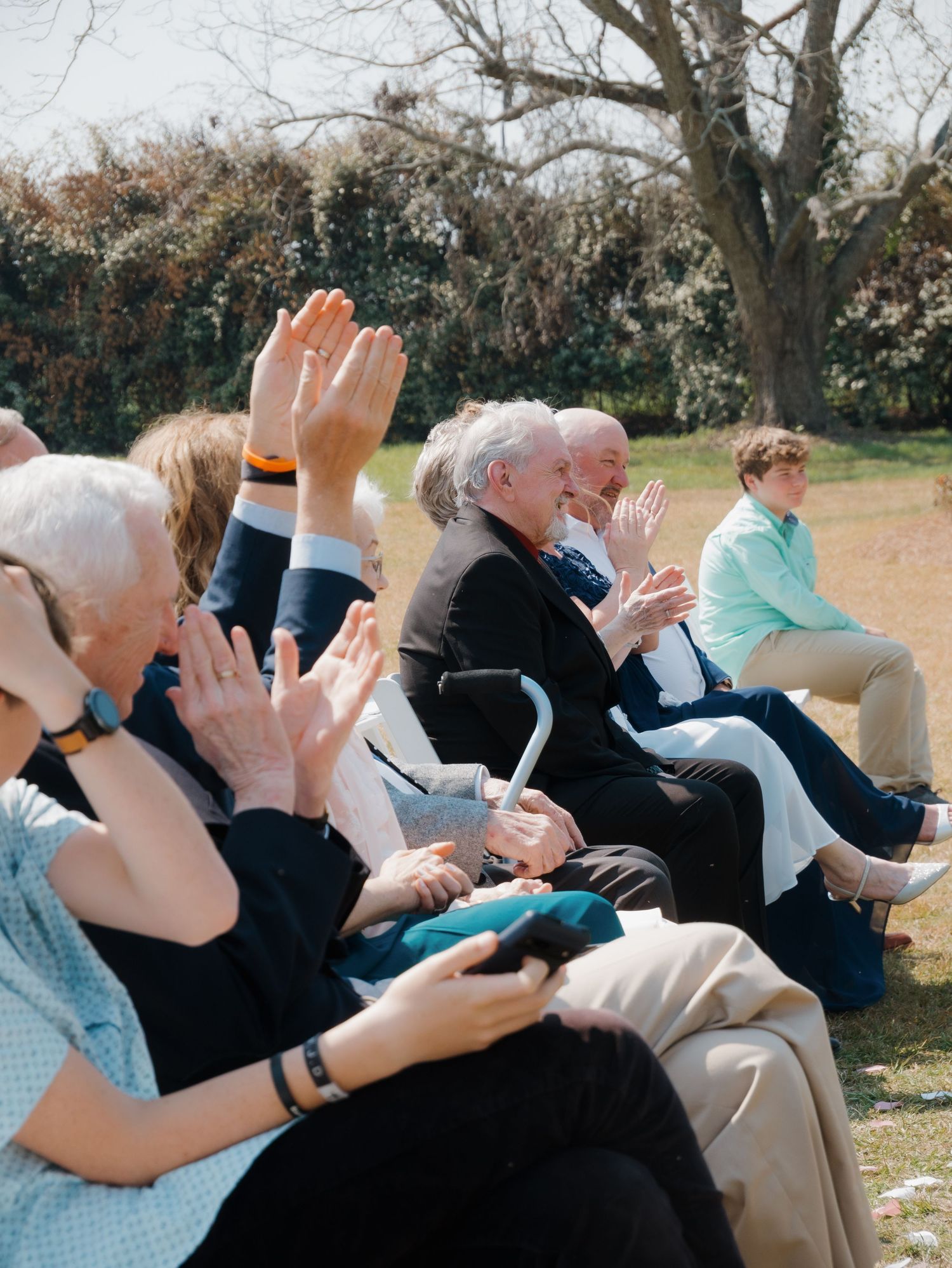 Wedding guests seated outdoors clapping during ceremony.