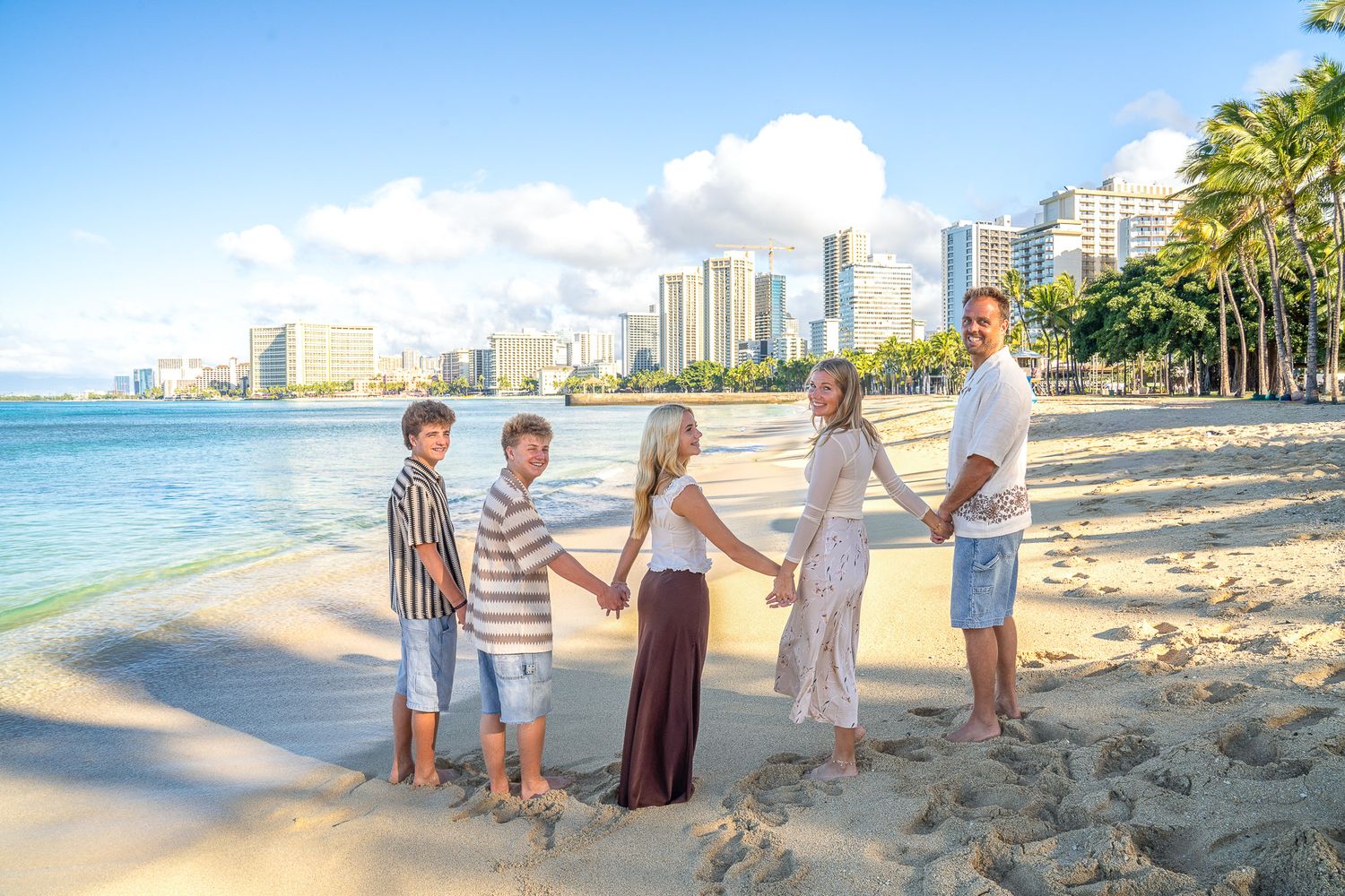 A group of people stand together on Waikiki Beach with palm trees and high-rise hotels in the background on a sunny day.