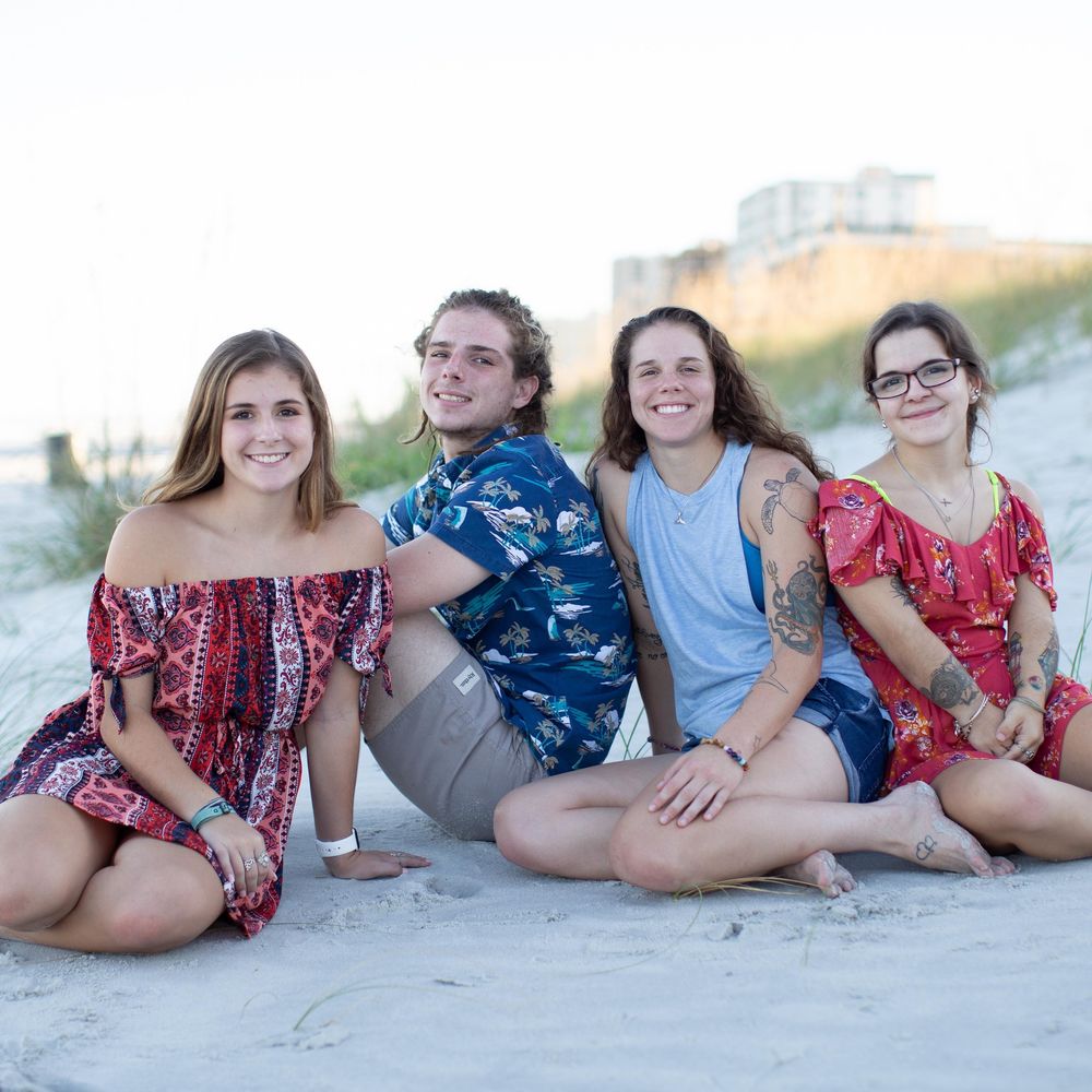 Four young adults smiling and sitting together on a sandy beach, wearing colorful summer outfits.