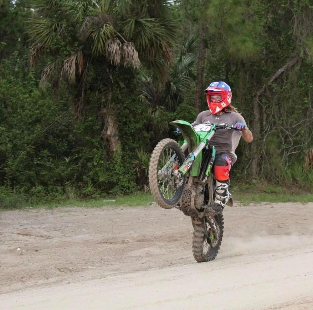 A motocross rider performs a wheelie on a green dirt bike along a sandy trail surrounded by lush palm trees.