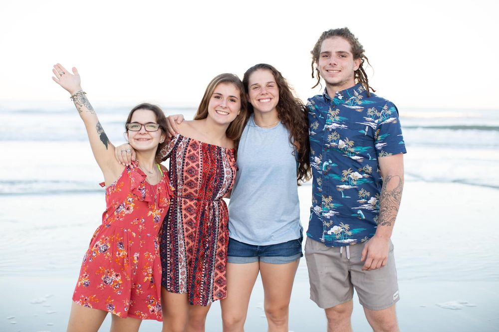 Four young friends smiling and posing together on a sunny beach, wearing colorful summer outfits.