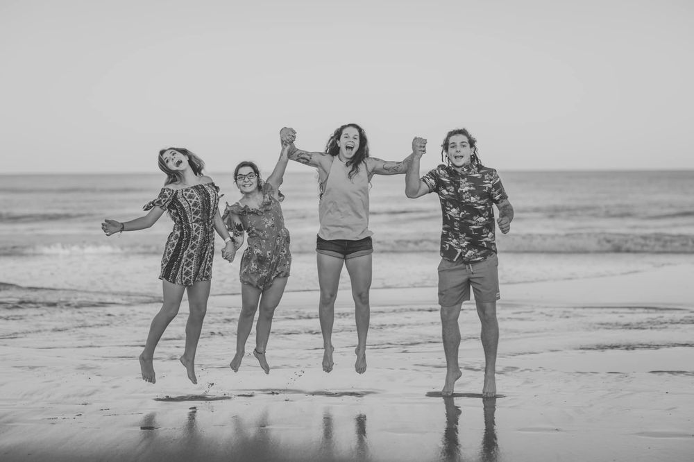 Four friends jumping joyfully on a beach in black and white photo, waves crashing behind them.