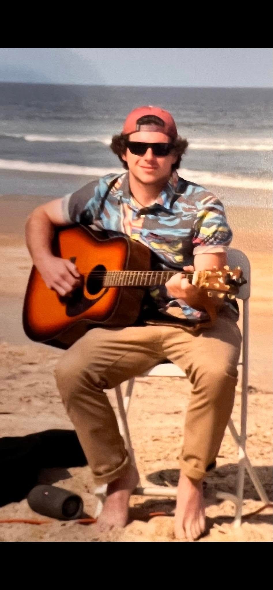 Young man playing acoustic guitar on beach, wearing hawaiian shirt, backwards cap, and sunglasses, seated in folding chair.