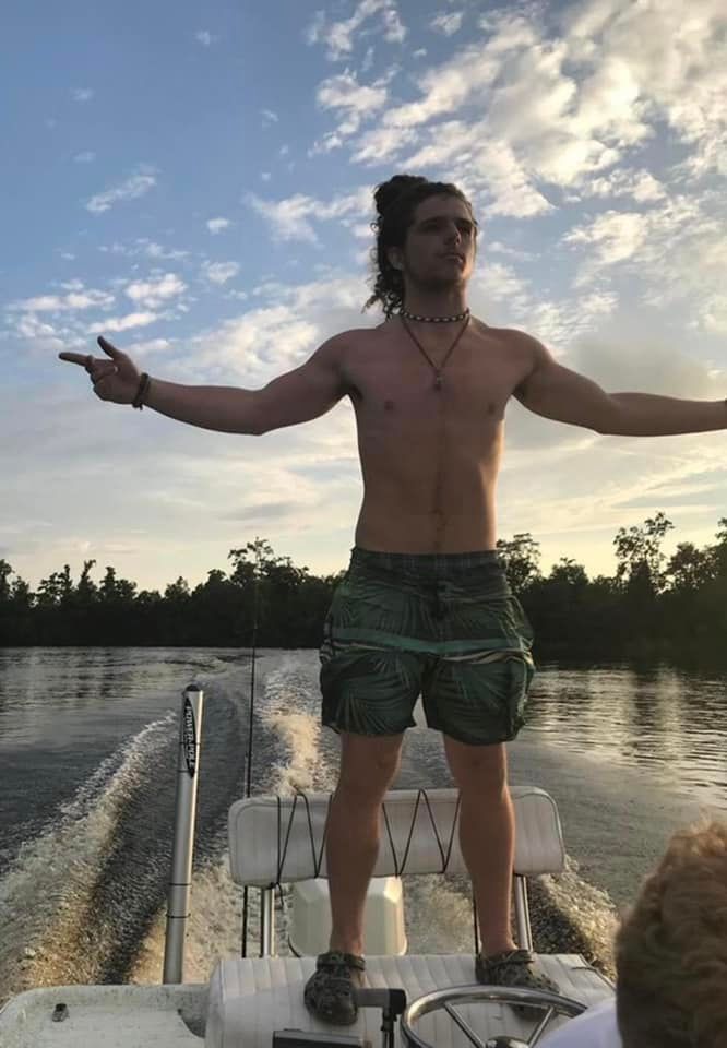 Shirtless young man with long hair standing on a boat with arms outstretched on a calm river at sunset.