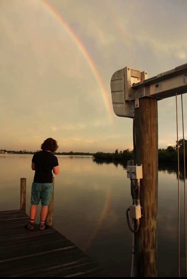 Child standing on a dock watching a stunning double rainbow reflect over calm lake waters at sunset.