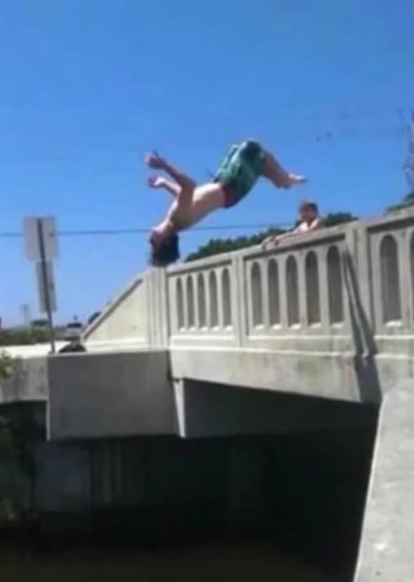 Person performing a backflip off a concrete bridge over a river on a sunny day.
