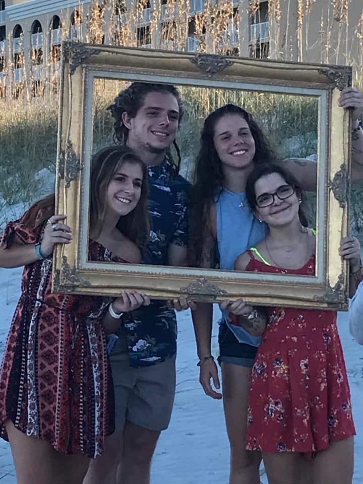 Four young friends posing with a decorative gold frame on a sandy beach at sunset.