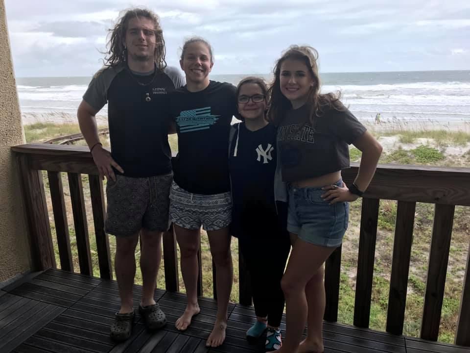 Four people smiling on a beach house deck with ocean waves and sandy shore visible in the background.