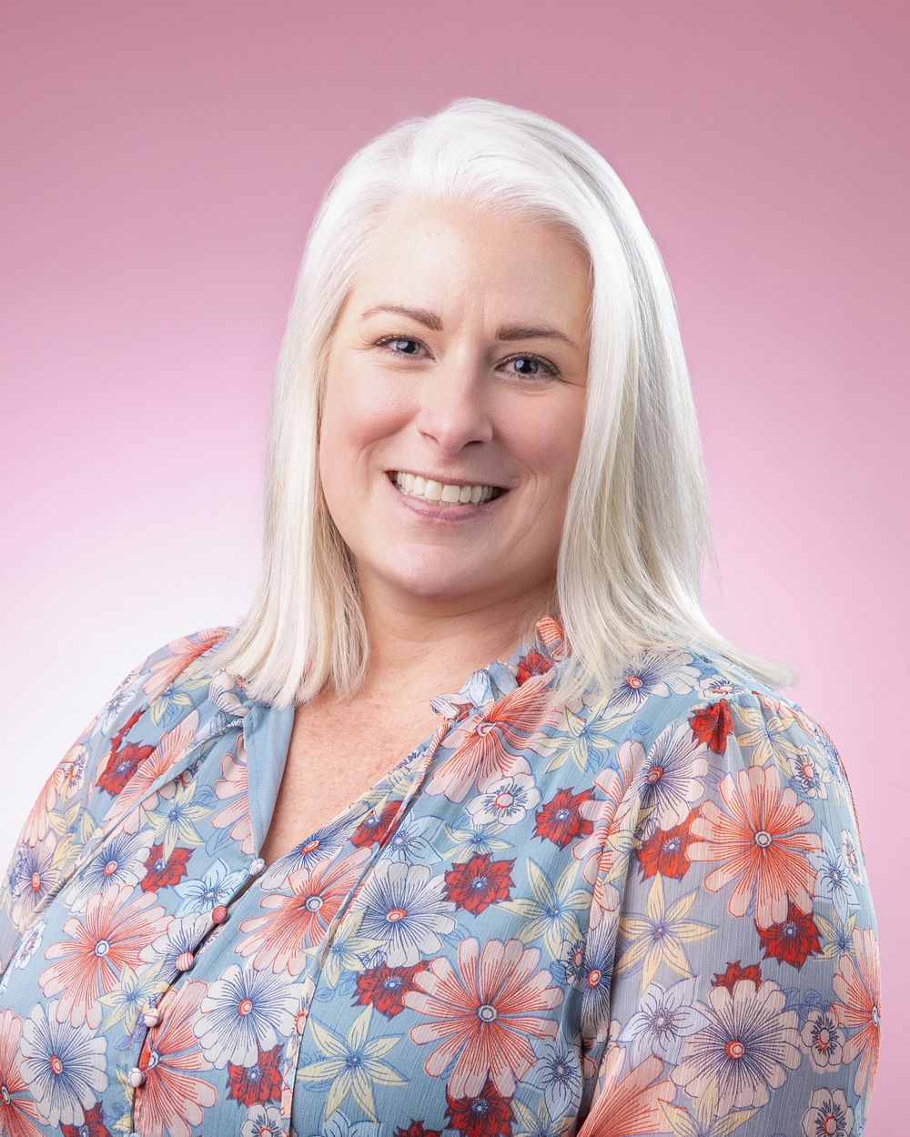 Professional headshot against pink background showing bright smile and platinum blonde hair wearing floral print blouse.