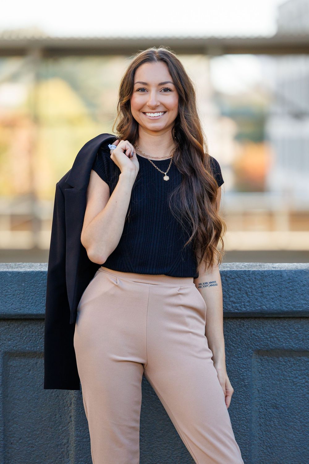 A person in business attire poses outdoors wearing a black top and beige pants against a dark wall.