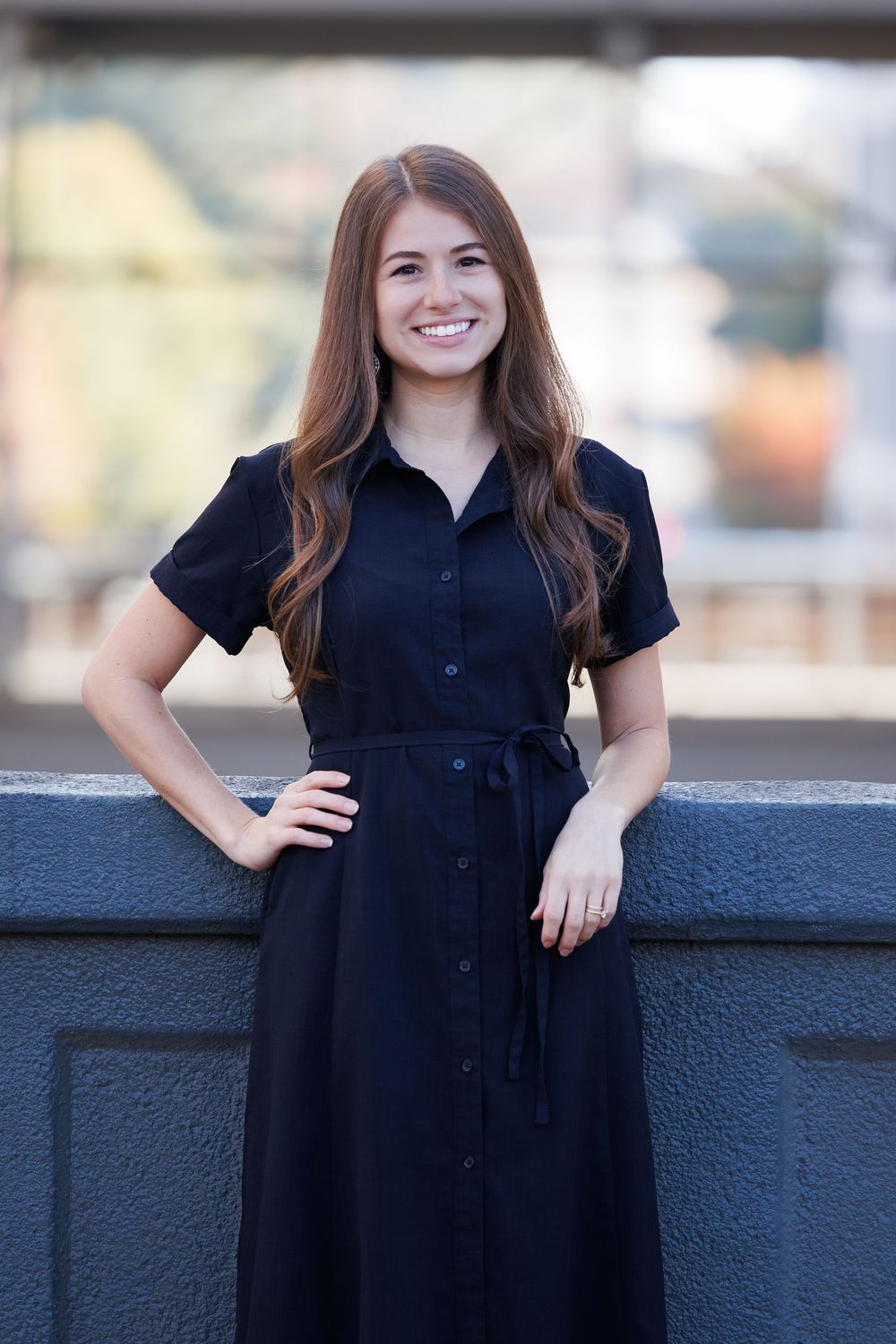 A person in a navy blue button-down dress with short sleeves poses outdoors while smiling and leaning against a wall.