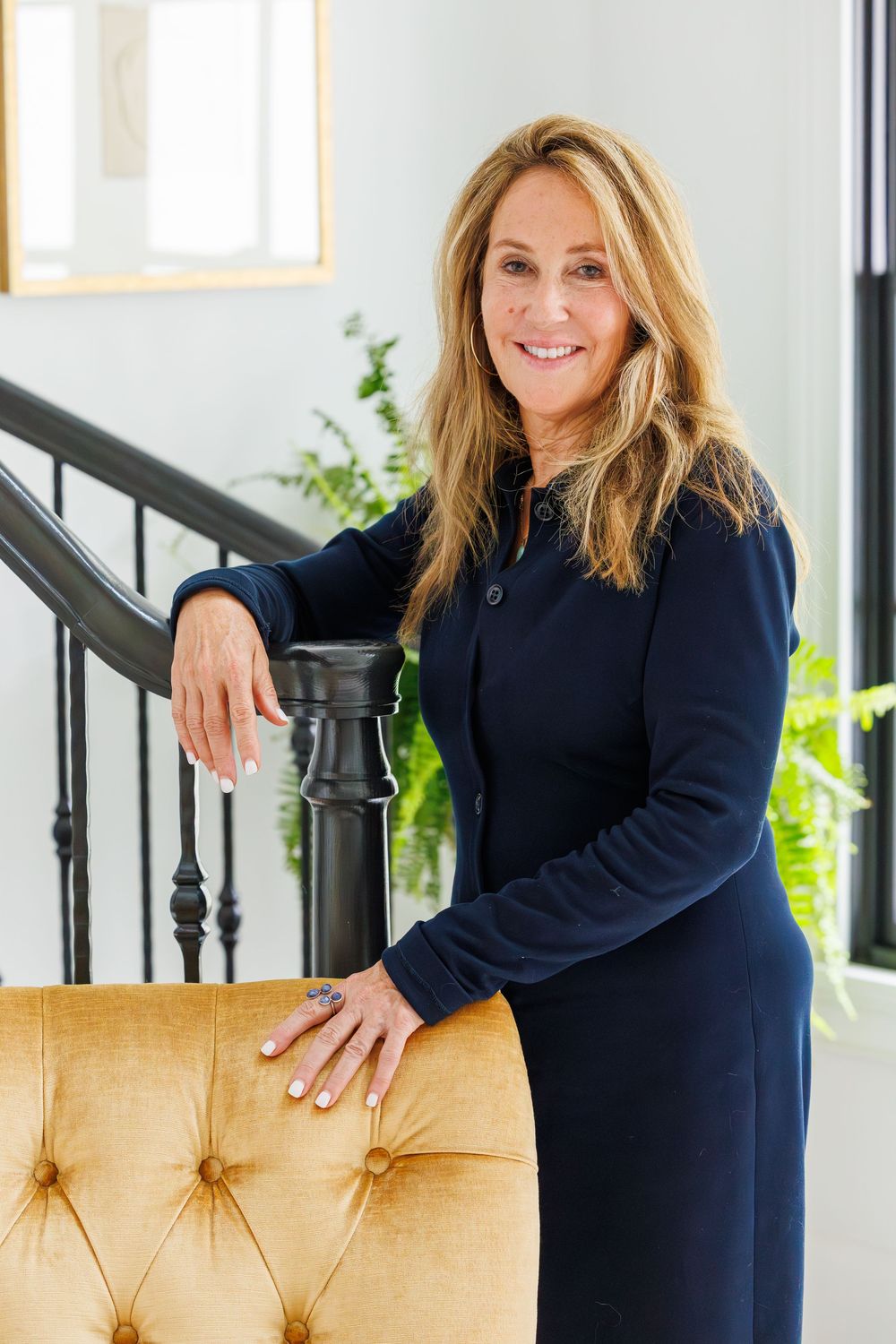 Professional woman in navy dress poses confidently next to a tufted chair and decorative stair railing.