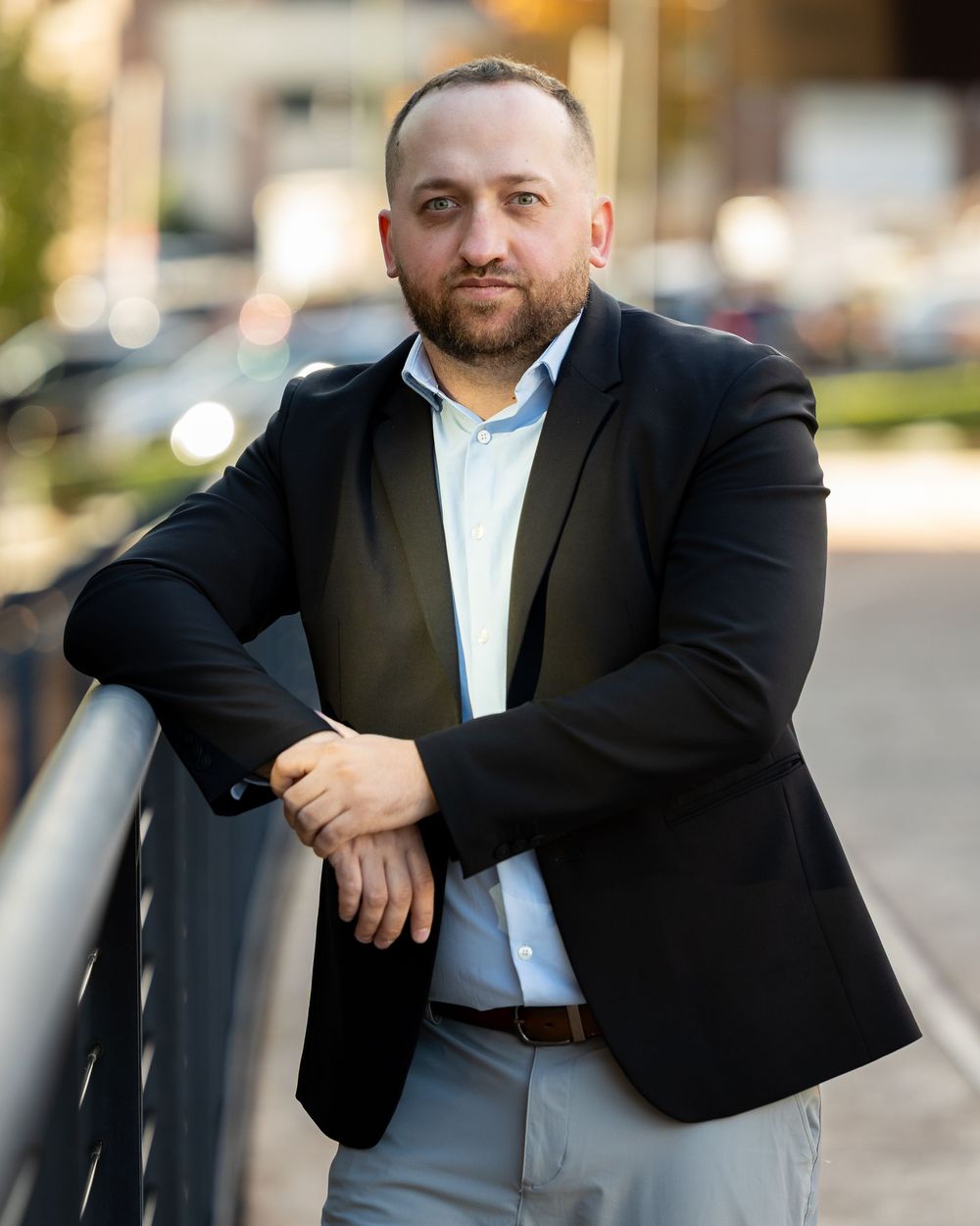 Professional portrait of someone in a black blazer and light blue dress shirt leaning on a railing outdoors.