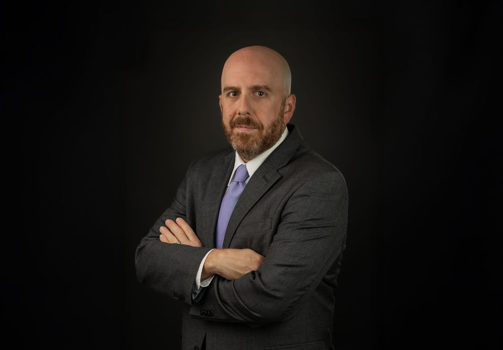 Professional business headshot series of someone in a dark suit and purple tie against a black background.