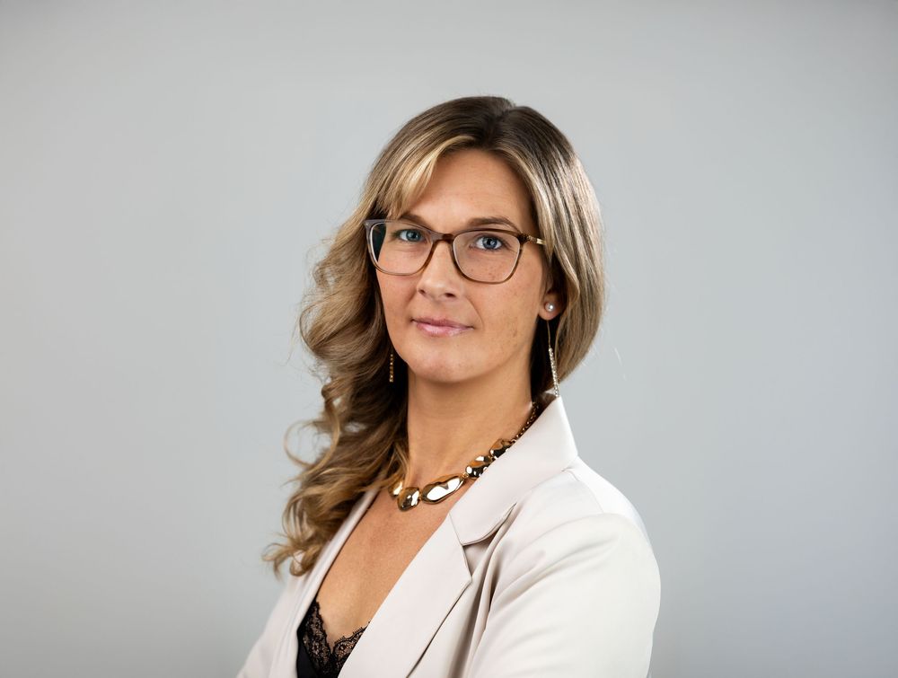 Professional woman in white blazer and glasses with wavy blonde hair poses for a corporate headshot portrait.