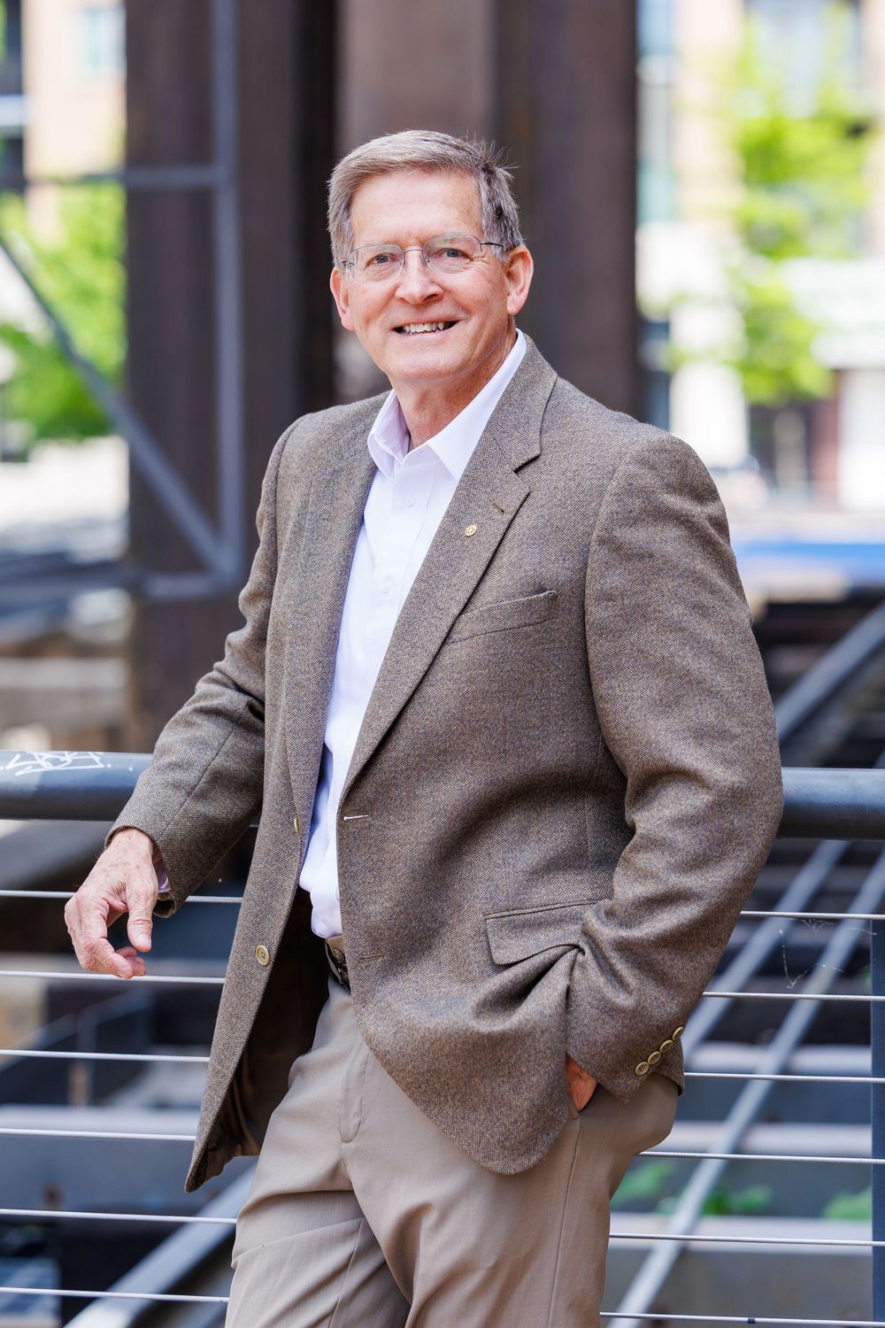 A person in a brown sport coat and khaki pants stands casually on outdoor steps with railing.