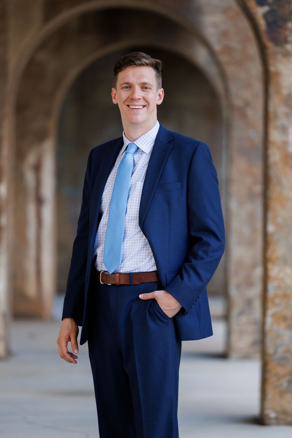 A person in a navy suit and light blue tie poses in front of stone archways in a series of professional portraits.