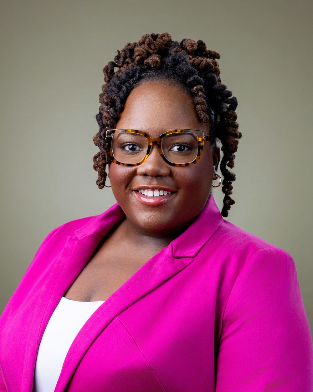 Professional headshot of someone wearing a bright pink blazer, glasses, and natural curled hairstyle against neutral background.