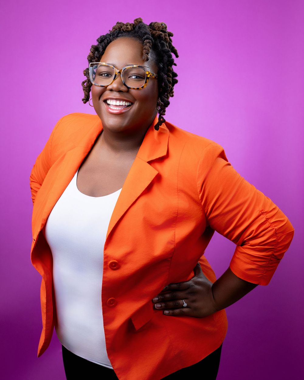 Professional portrait against purple background showing bright orange blazer, white top, glasses and a confident pose.