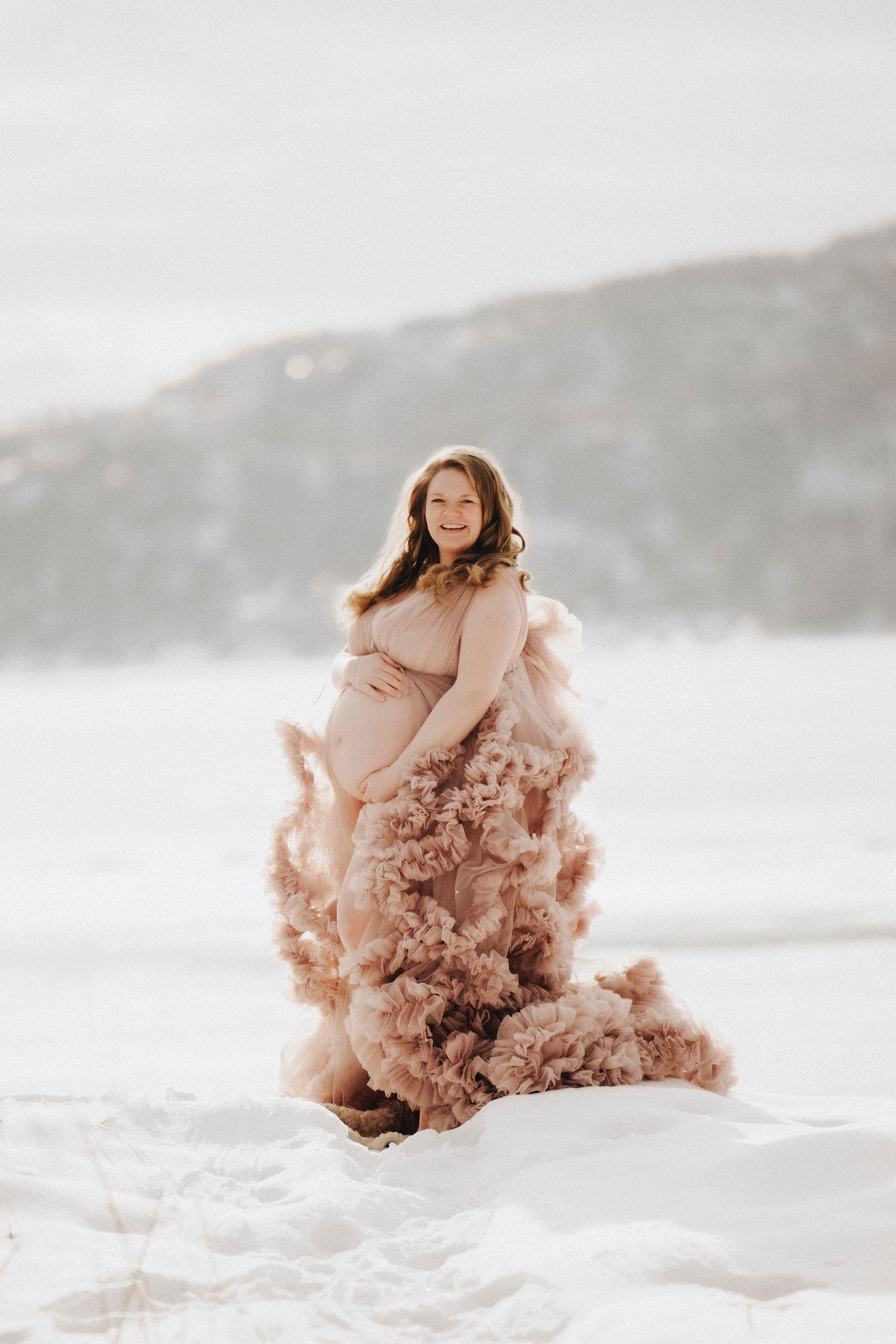 A dreamy beach scene with a flowing blush pink tulle dress against white sand and misty coastal mountains.