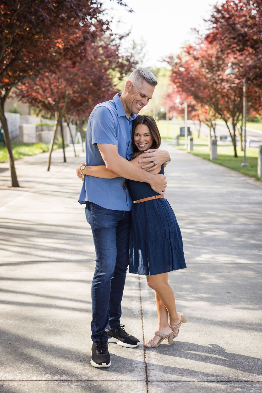 A couple in blue and navy outfits embrace lovingly on a tree-lined pathway during an outdoor photo session.