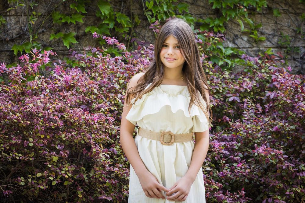 A person in a cream-colored dress poses in front of blooming pink flowers and green foliage in a garden setting.